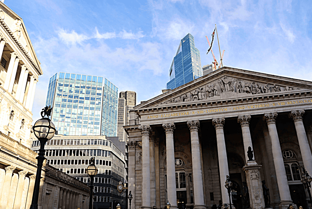 Historic Bank of England building with classical columns in the foreground, surrounded by modern glass skyscrapers in the City of London under a blue sky.