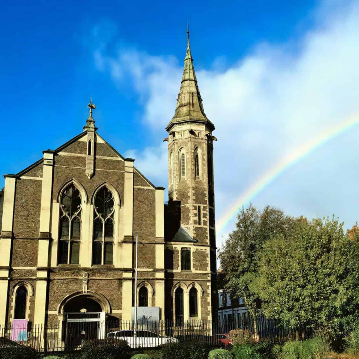 Traditional stone church with tall steeple and a rainbow in the sky beside it on a sunny day.