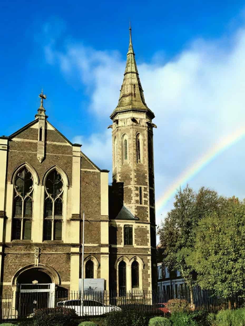 Traditional stone church with tall steeple and a rainbow in the sky beside it on a sunny day.
