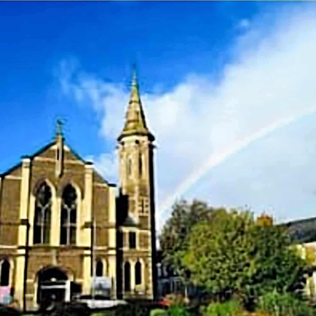 Historic stone church with tall spire under a bright blue sky and a rainbow arching overhead, with nearby houses and trees in the foreground.