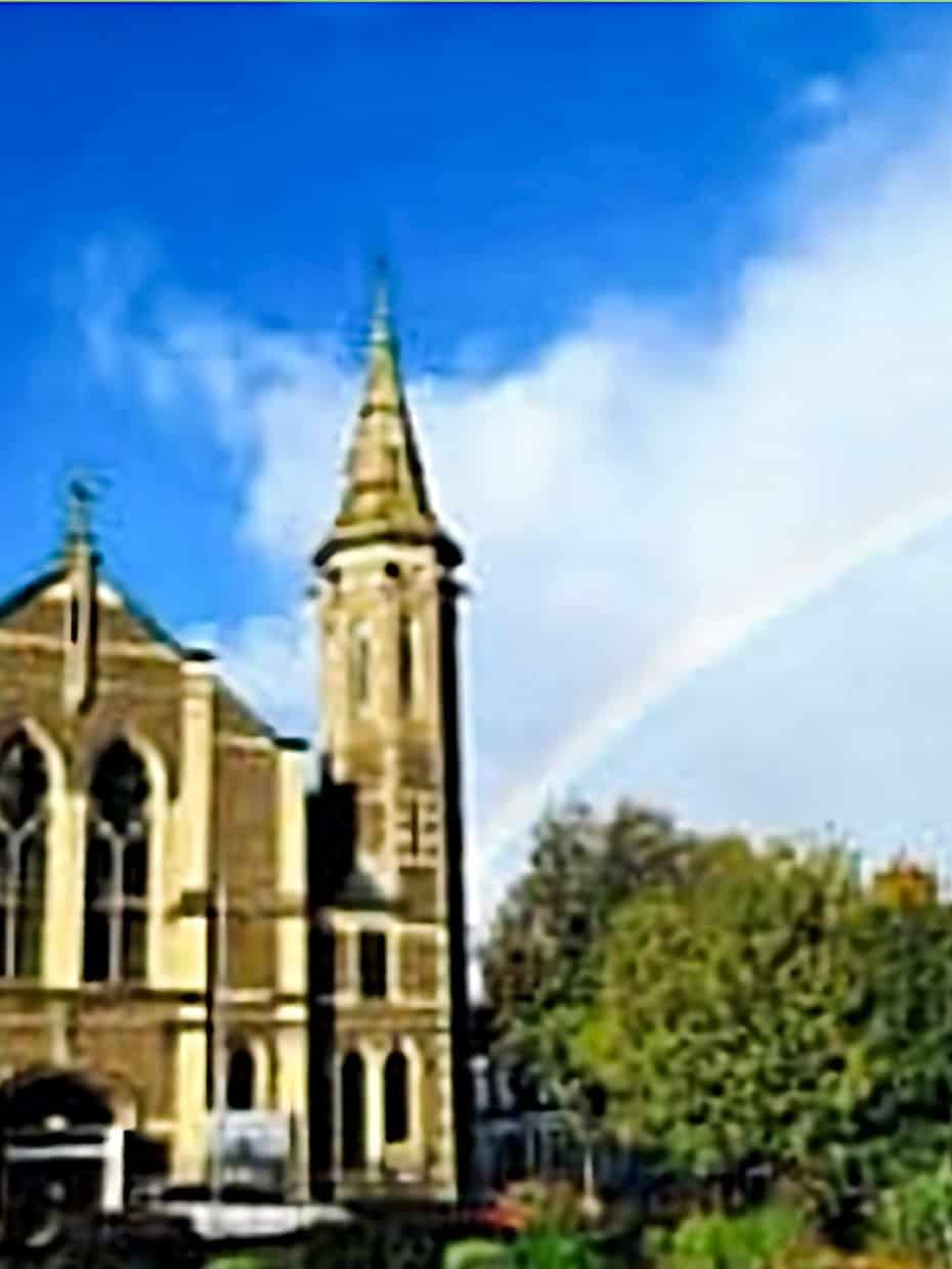 Historic stone church with tall spire under a bright blue sky and a rainbow arching overhead, with nearby houses and trees in the foreground.