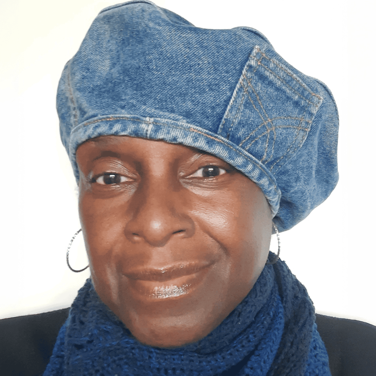 Smiling woman wearing a blue denim beret and scarf, facing the camera against a plain light background.