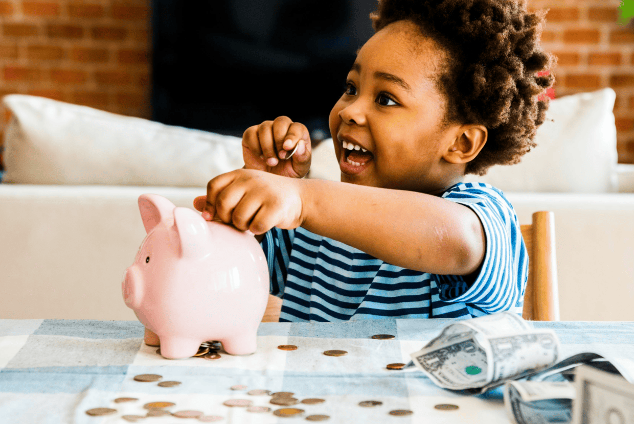 Young child smiling while placing coins into a pink piggy bank, with loose change on the table, representing early saving habits and financial learning.