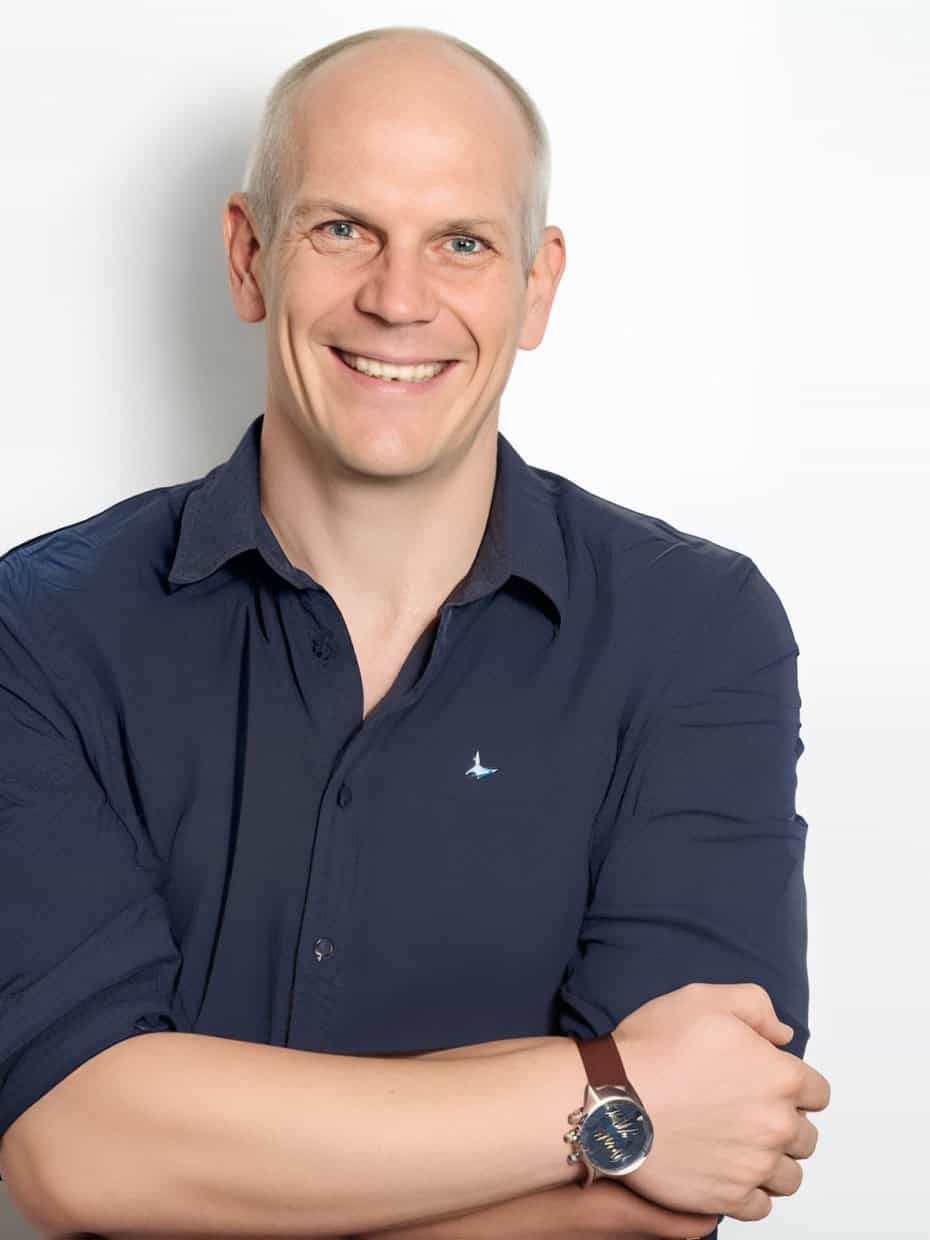Smiling man with folded arms wearing a dark blue shirt and watch, standing against a plain white background.