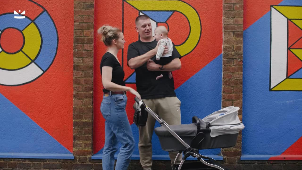 Parents standing with a baby and pram in front of a bright geometric mural wall.