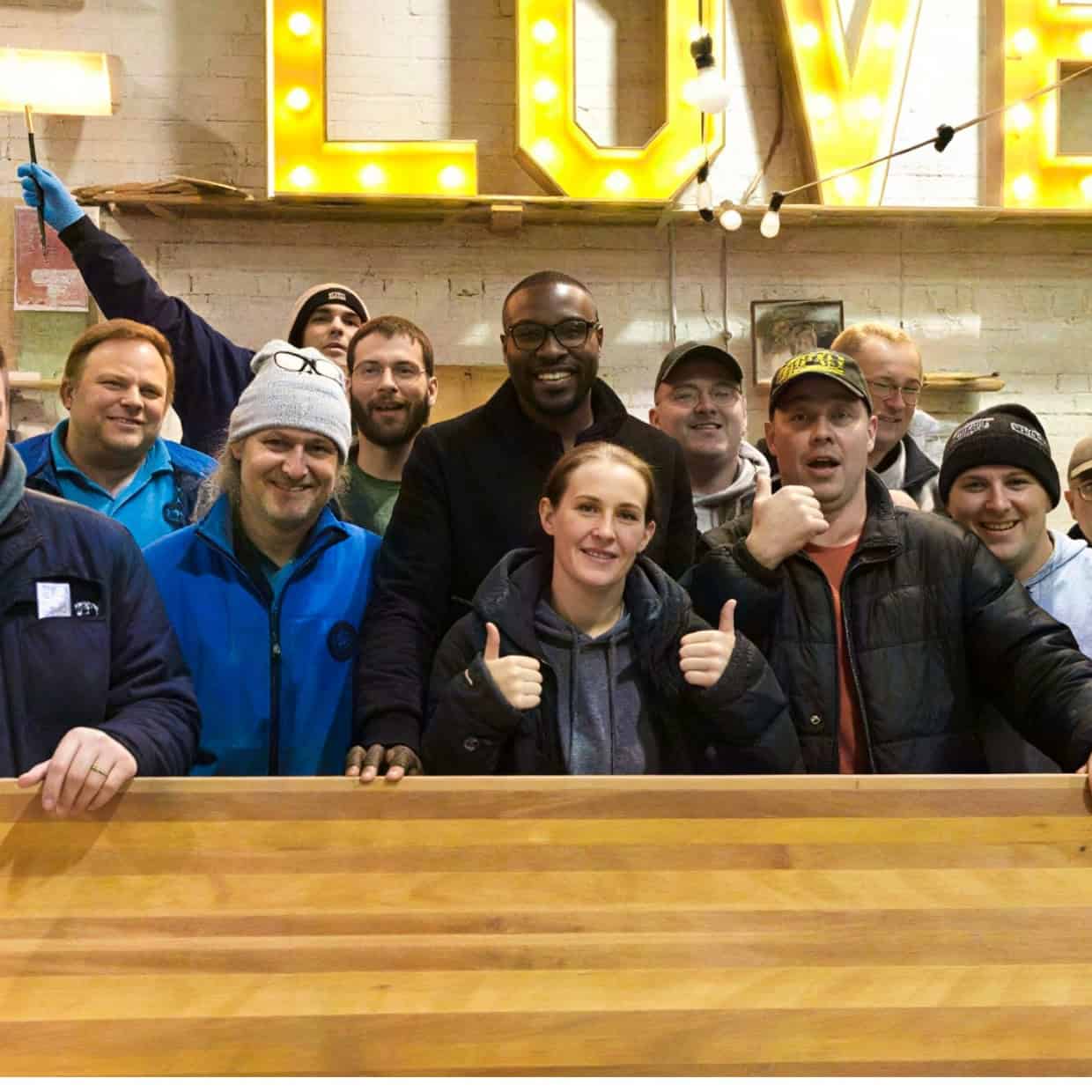 Group of people from Handcrafted Projects standing together in a workshop in Gateshead with a large wooden piece and illuminated sign behind them.
