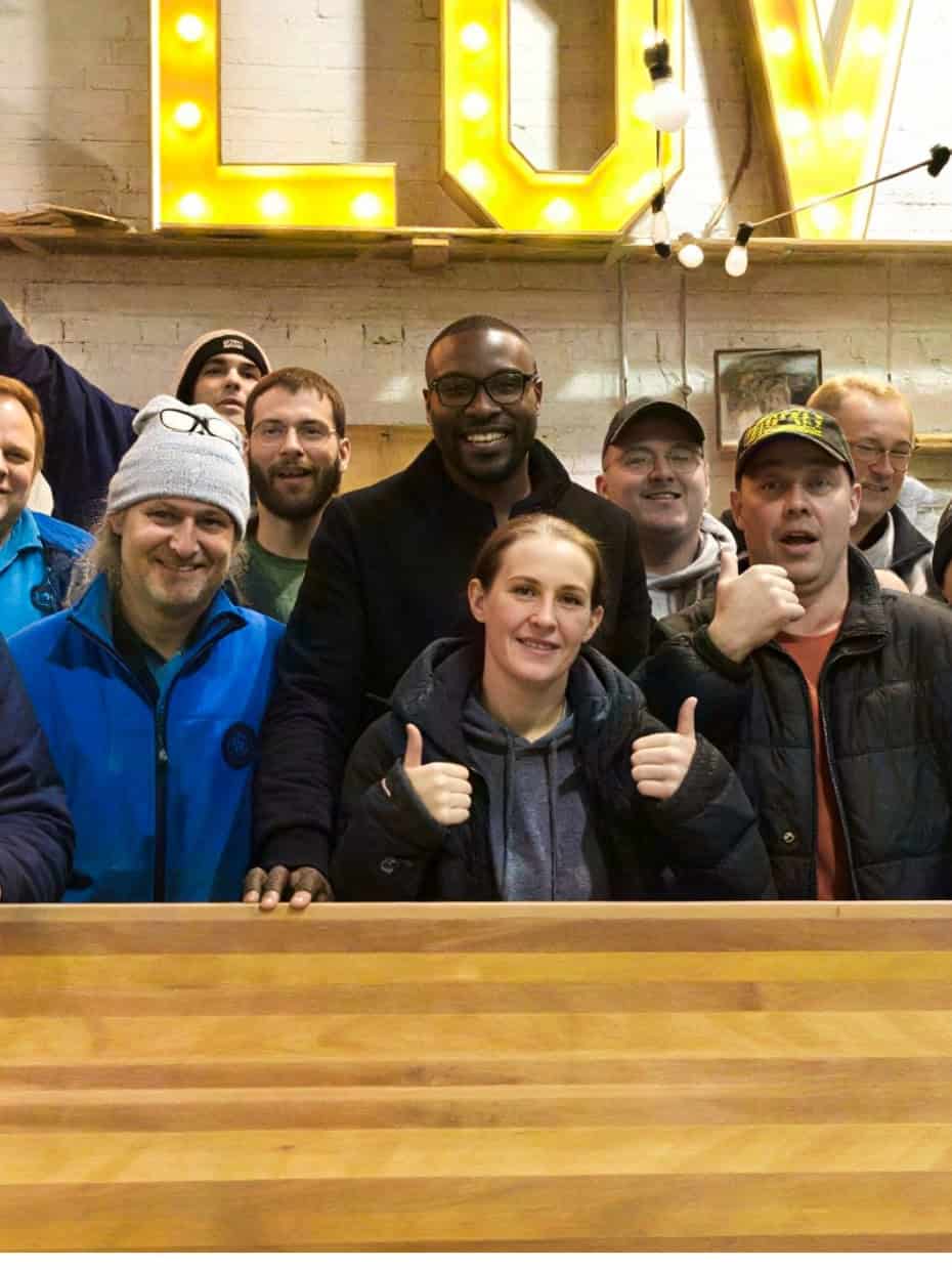 Group of people from Handcrafted Projects standing together in a workshop in Gateshead with a large wooden piece and illuminated sign behind them.