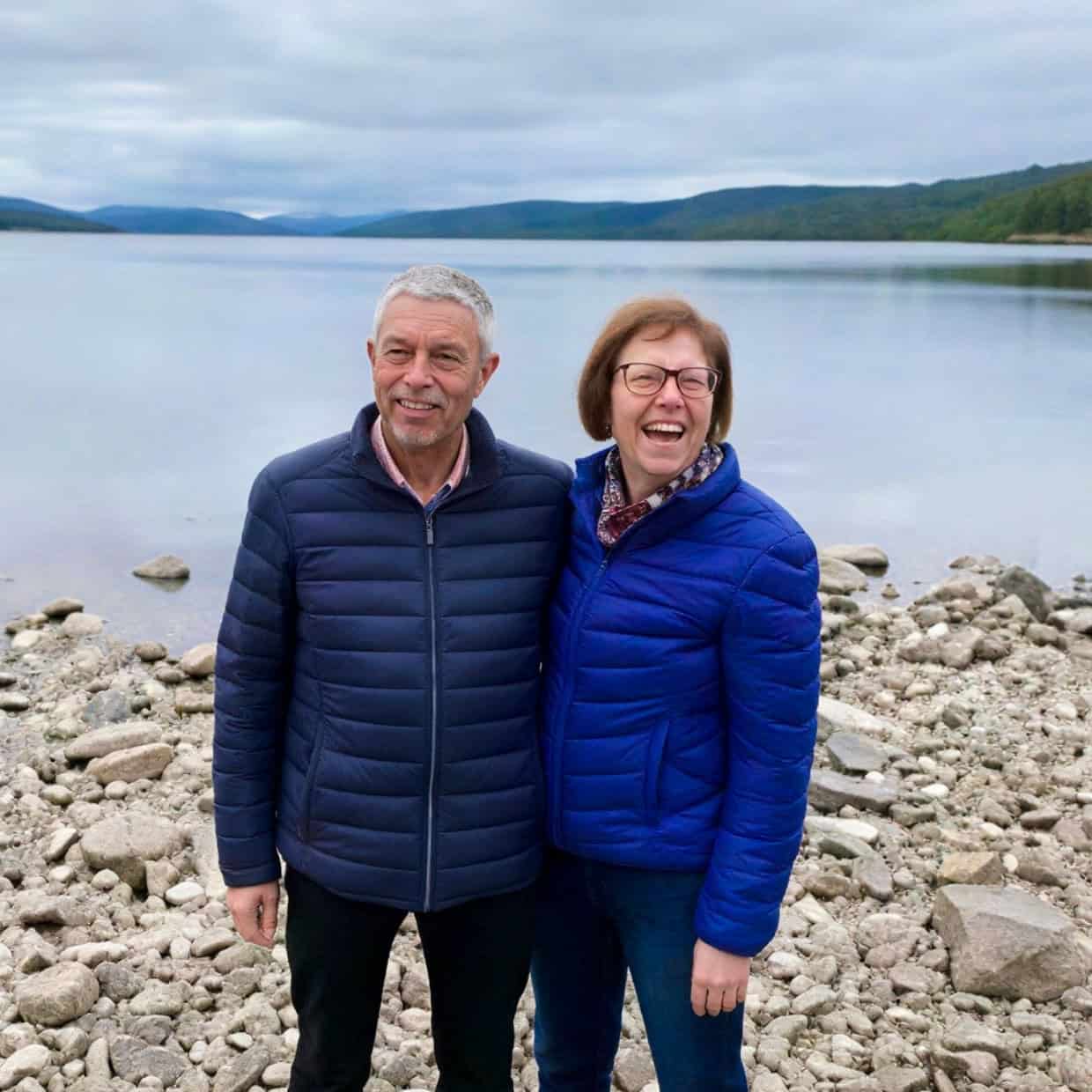 Smiling couple standing on a rocky lakeshore with calm water and mountains behind them.