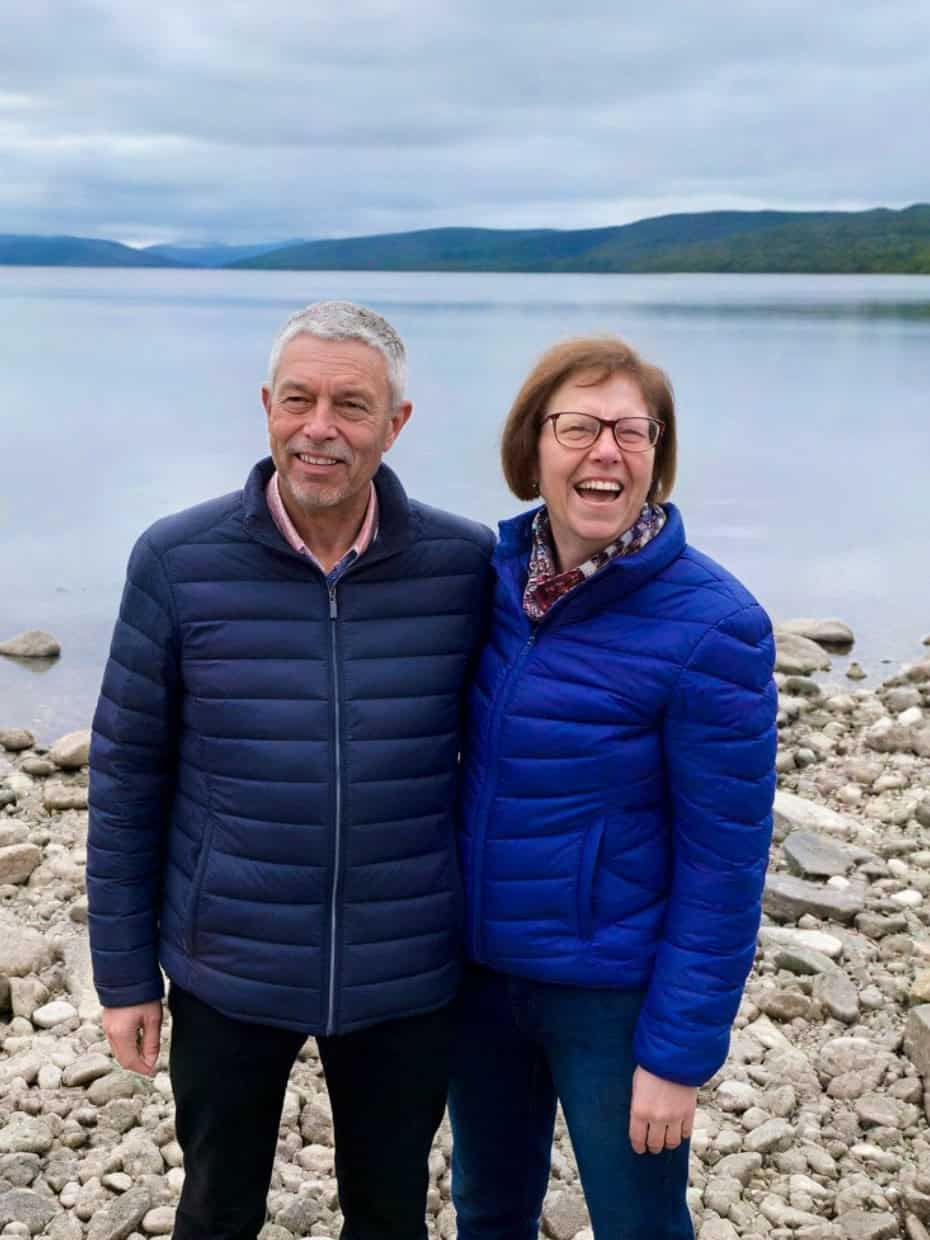 Smiling couple standing on a rocky lakeshore with calm water and mountains behind them.