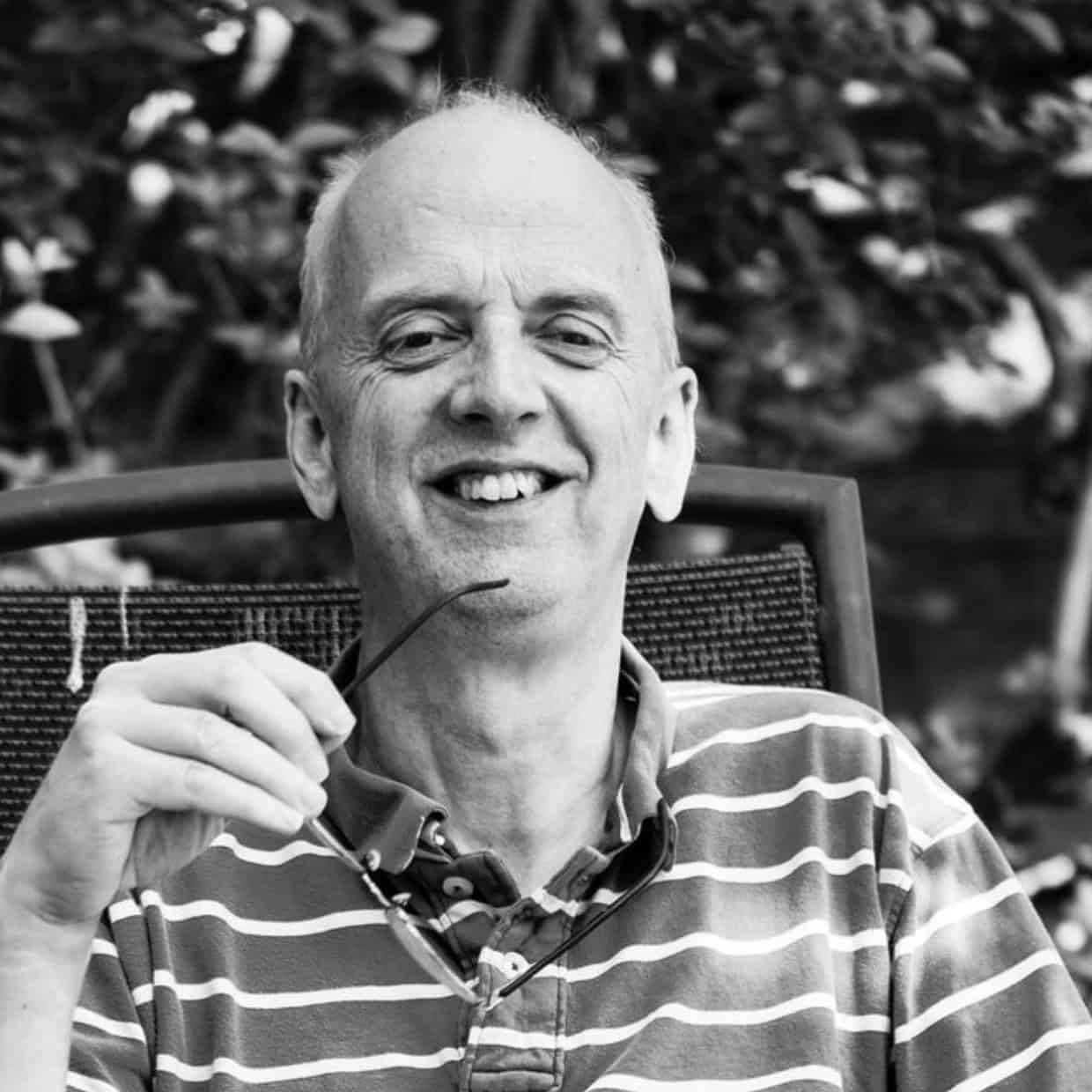 Jeremy Marshall smiling while holding glasses, seated outdoors with trees in the background.
