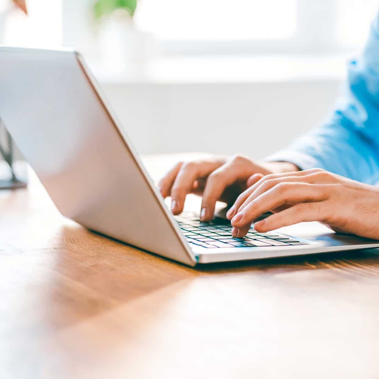 Close up of hands typing on a laptop keyboard on a desk in a bright workspace.