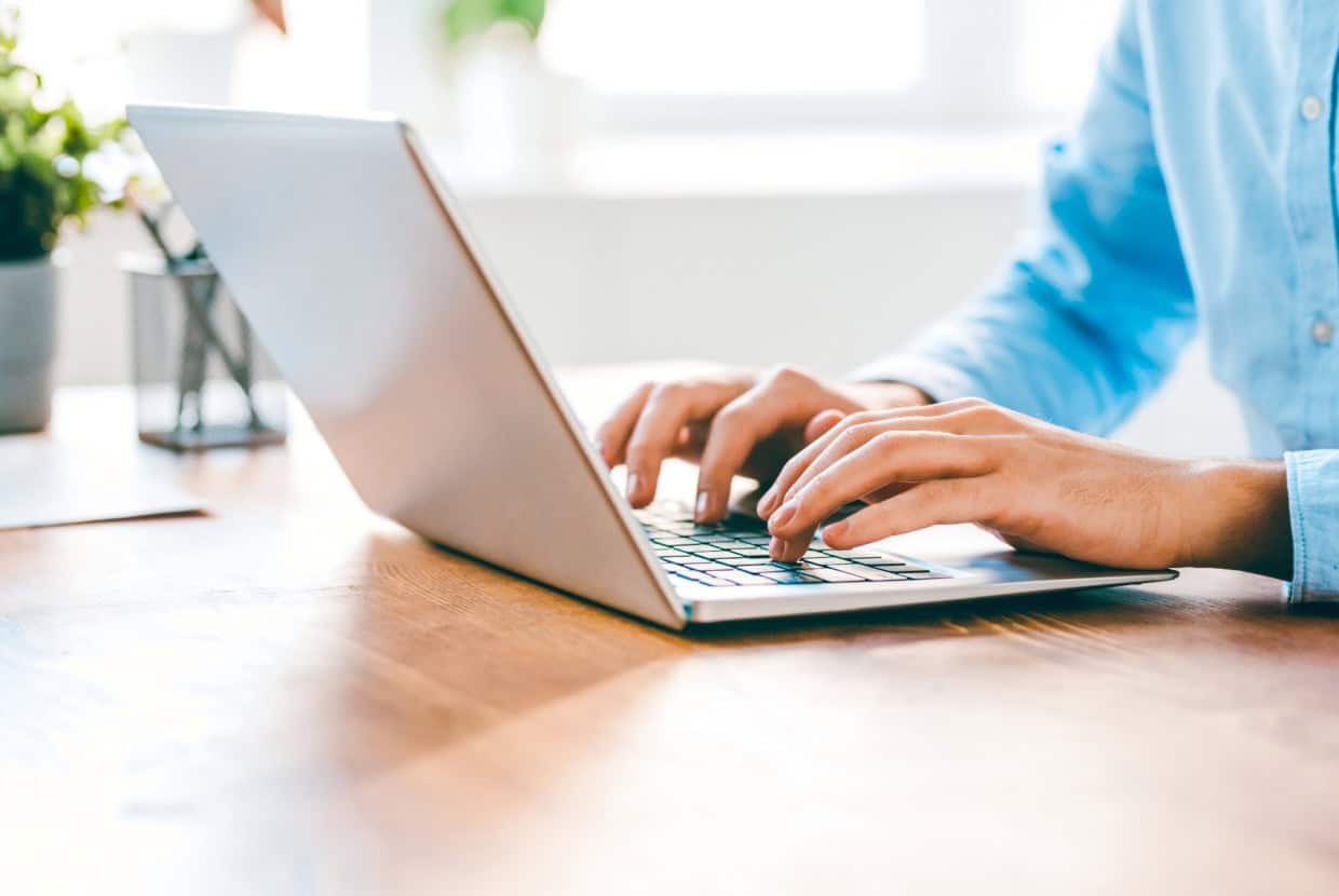 Close up of hands typing on a laptop keyboard on a desk in a bright workspace.