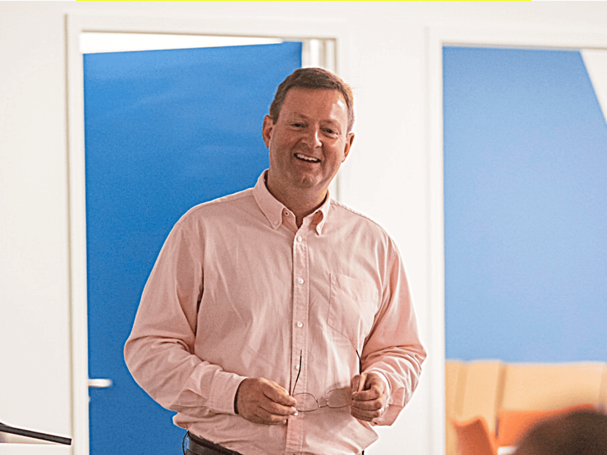 Paul Houghton smiling while speaking indoors, holding glasses in his hands with a bright background behind him.