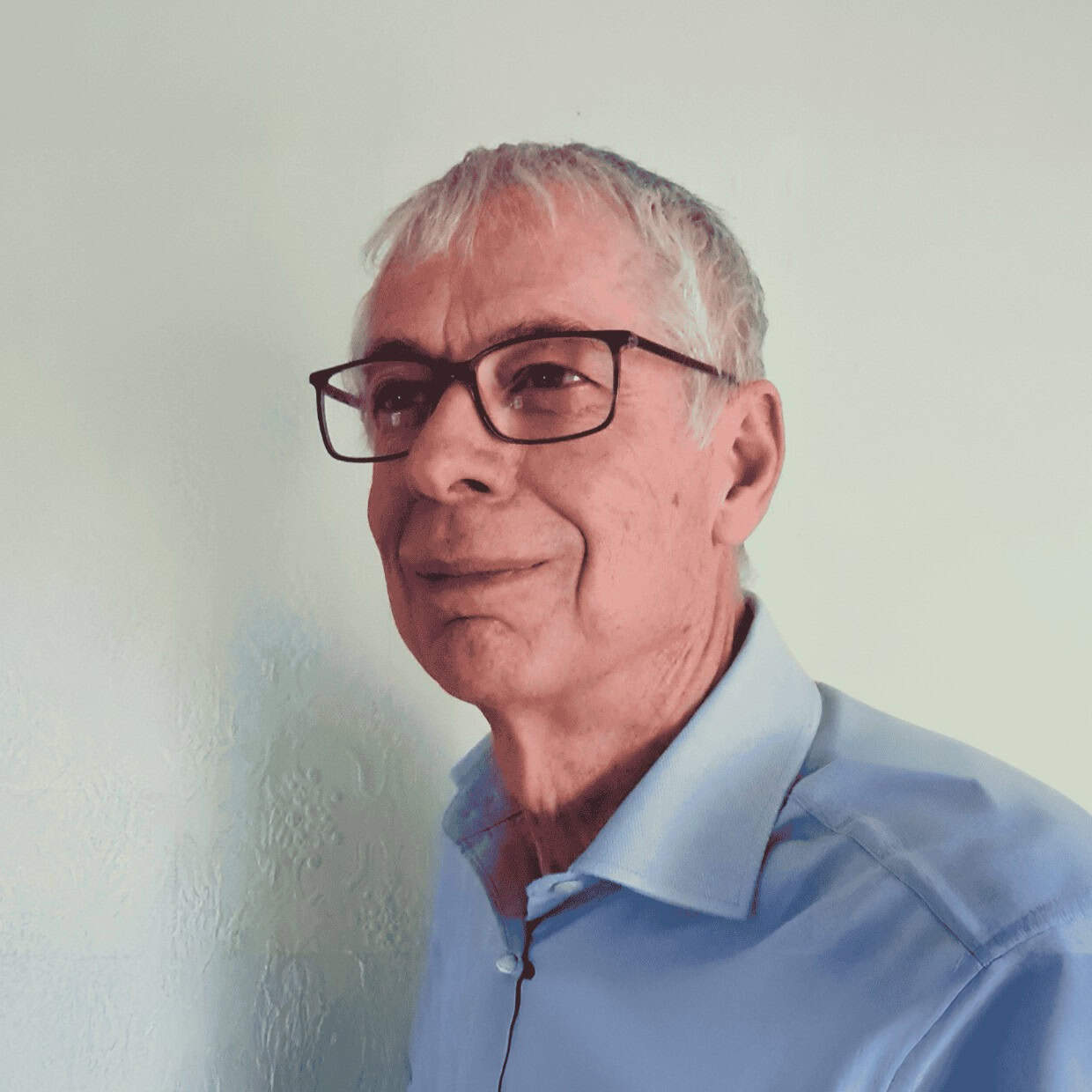 A man with grey hair and glasses wearing a light blue shirt against a plain background.