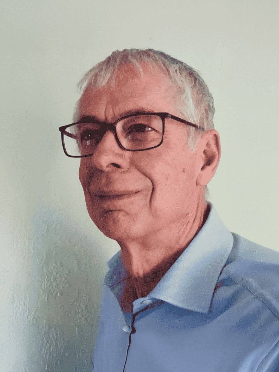 A man with grey hair and glasses wearing a light blue shirt against a plain background.