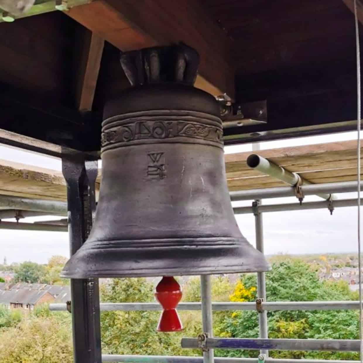 Large church bell hanging in a tower with view over rooftops and trees in the background.