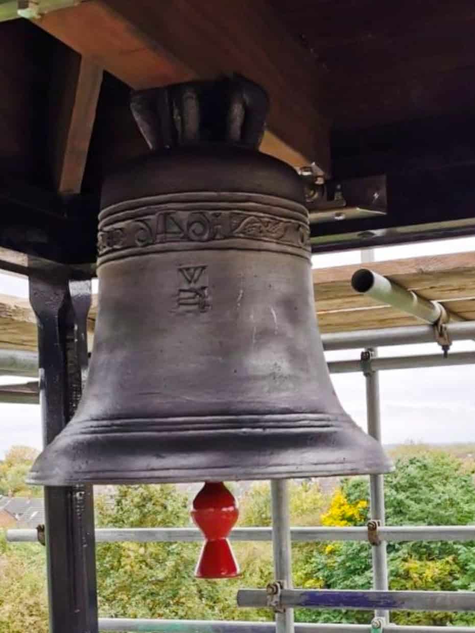 Large church bell hanging in a tower with view over rooftops and trees in the background.