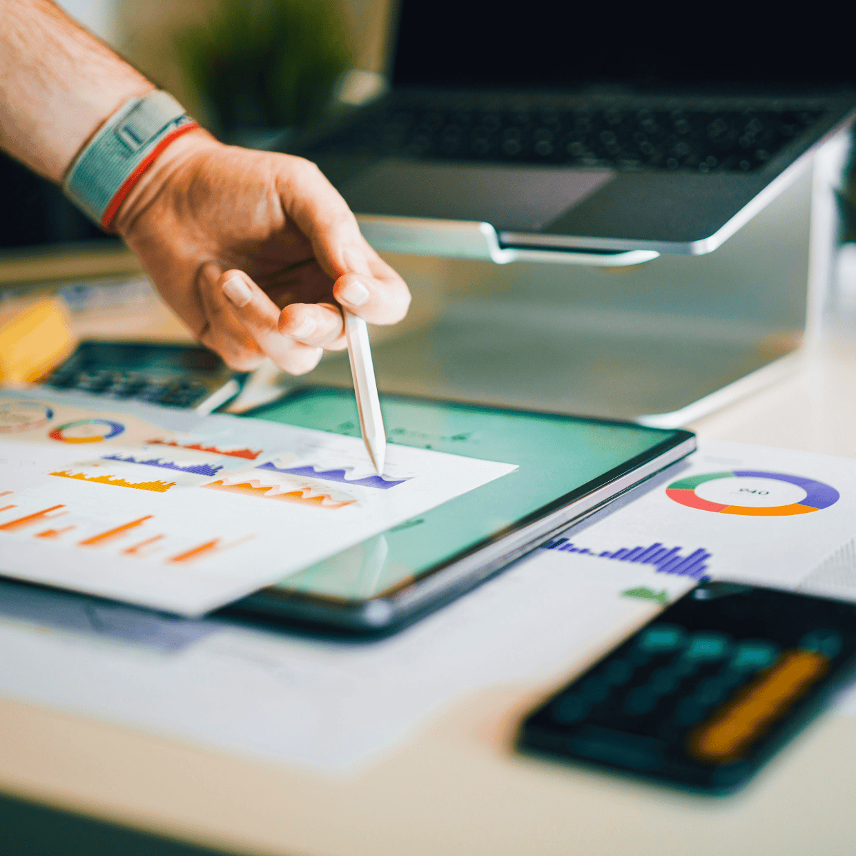 Hand using a stylus on a tablet displaying charts and graphs, with printed financial reports, calculator, and laptop on a desk.