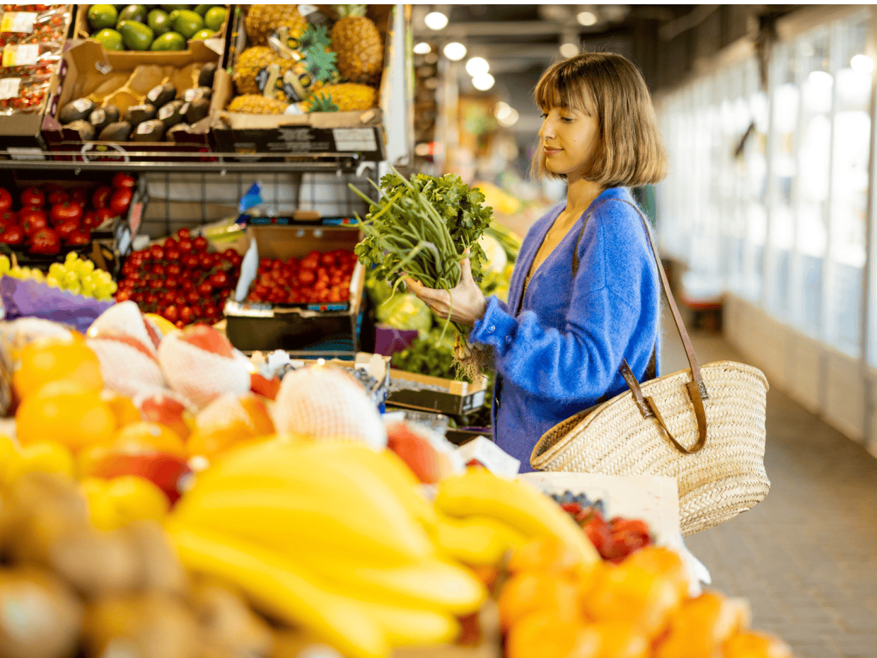 Woman selecting fresh vegetables at a market stall with fruit and produce displayed around her.