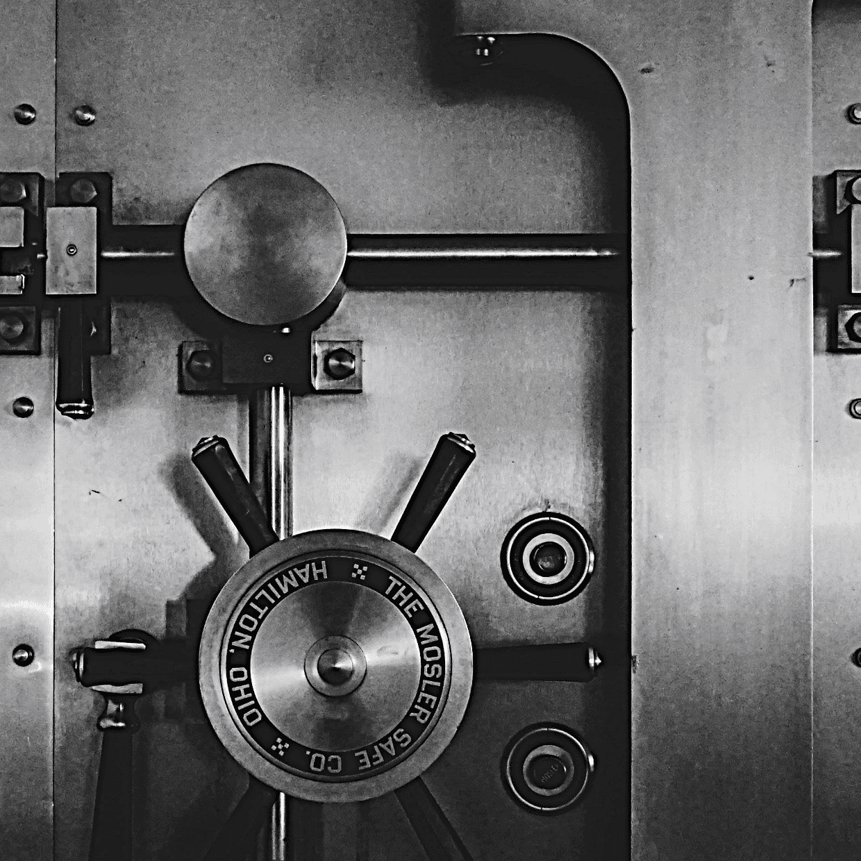 Close up of a heavy steel bank vault door with locking mechanism and wheel handle in black and white.