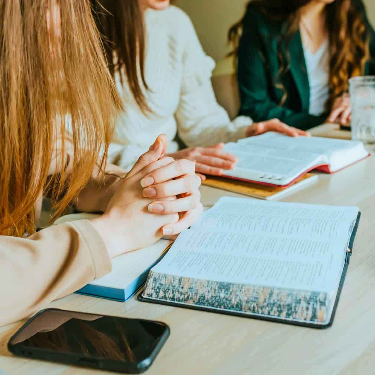 Group of people gathered around a table with open Bibles, studying scripture together, one person clasping their hands in prayer.