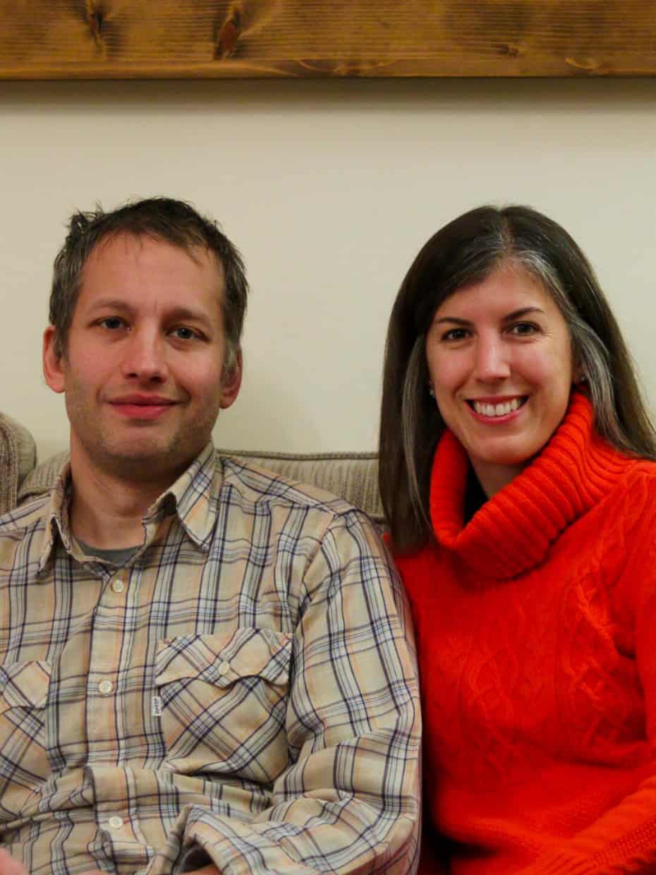 Man in a checked shirt and woman in a bright orange jumper sitting together on a beige sofa, smiling at the camera.