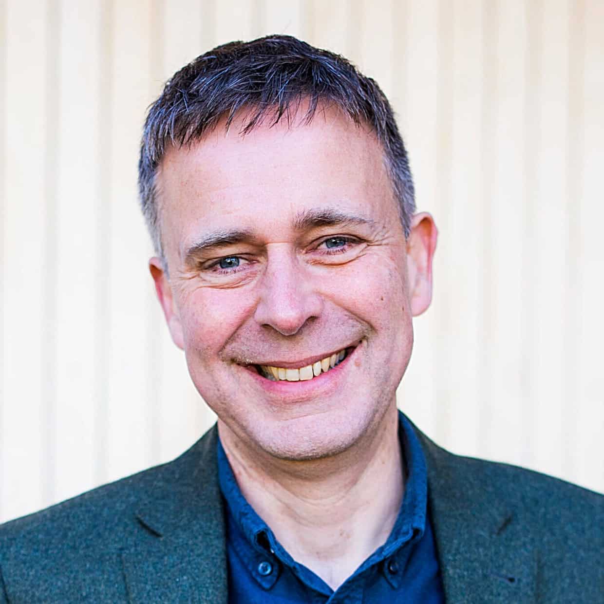 Smiling man wearing a dark green blazer and blue shirt, standing in front of a light vertical panel background.