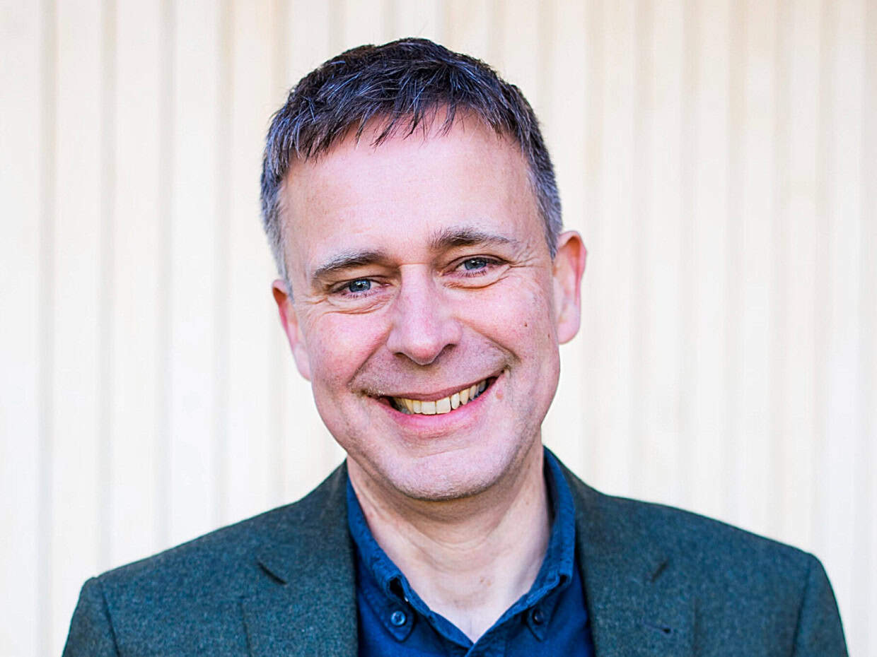Smiling man wearing a dark green blazer and blue shirt, standing in front of a light vertical panel background.