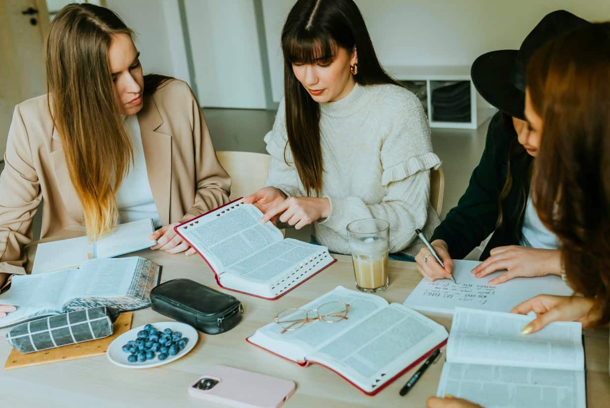 Group of young women sitting around a table studying together with open Bibles and notebooks.