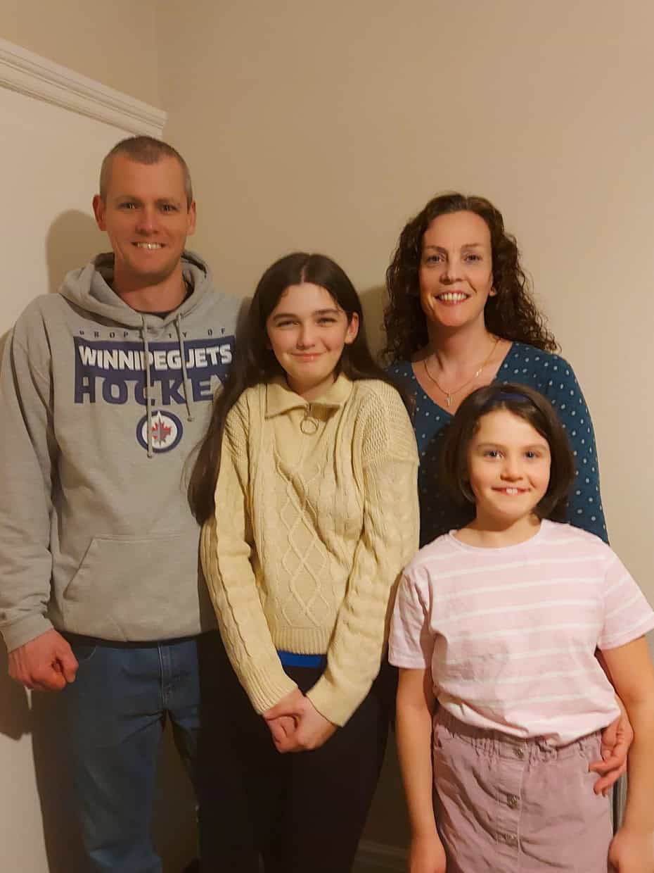 Smiling family of four standing together indoors, with parents behind and two daughters in front near a light-coloured wall and sofa.