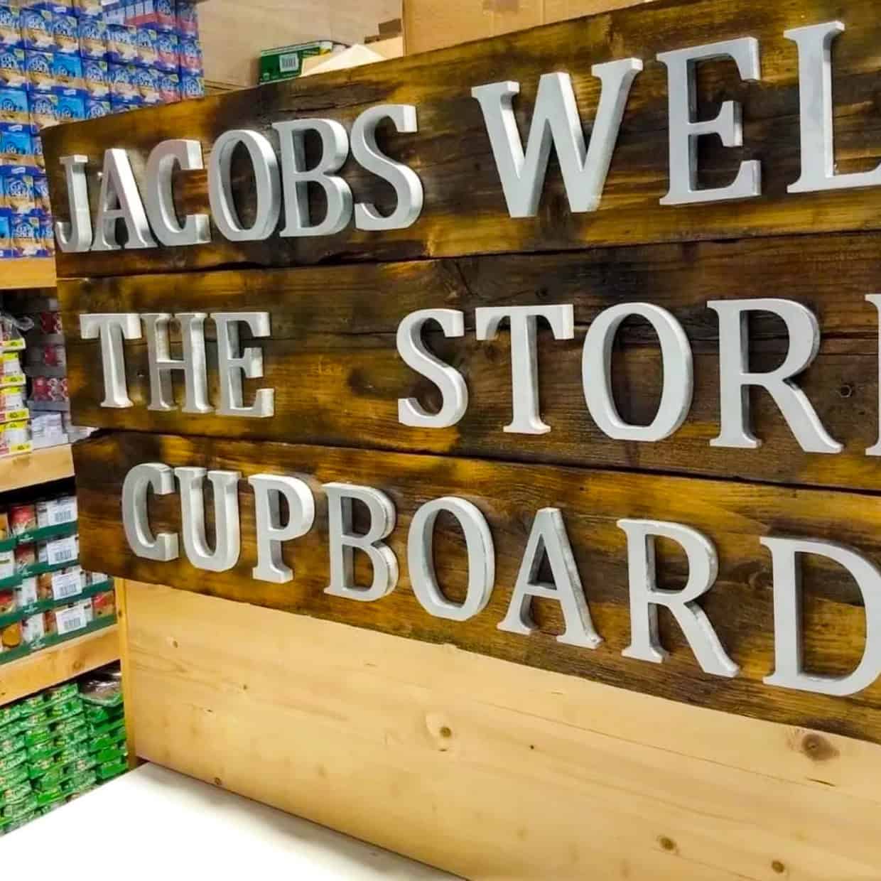 Wooden sign reading “Jacobs Well The Store Cupboard” displayed inside a food storage area with shelves of packaged goods in the background.