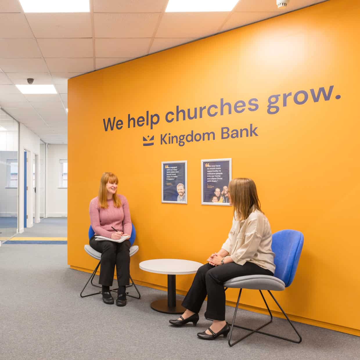 Two women talking in front of an orange wall with the words "We help churches grow. Kingdom Bank" displayed.