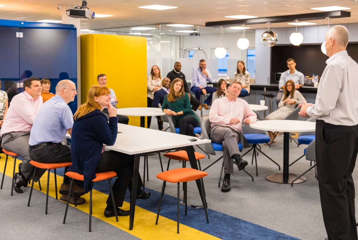 A group of employees listening to a speaker during a team meeting in a modern office.
