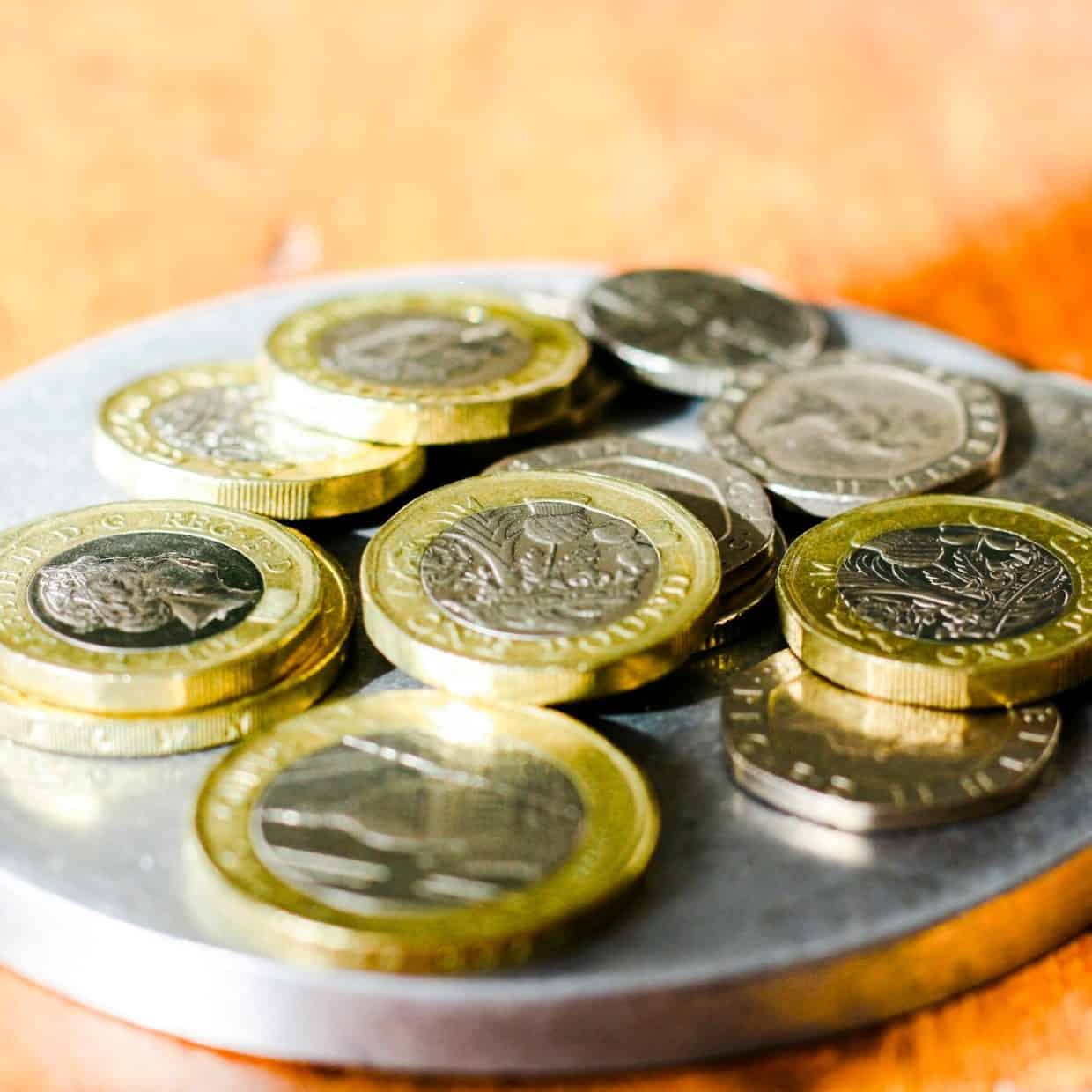 Close-up of British pound coins and small change stacked on a round metal tray on a wooden surface.