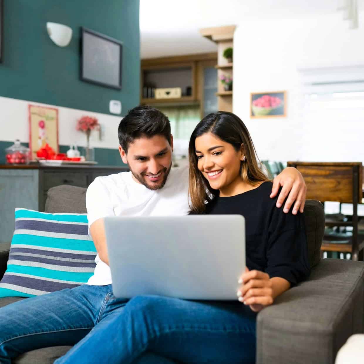 Smiling couple sitting on a sofa at home, looking at a laptop together.