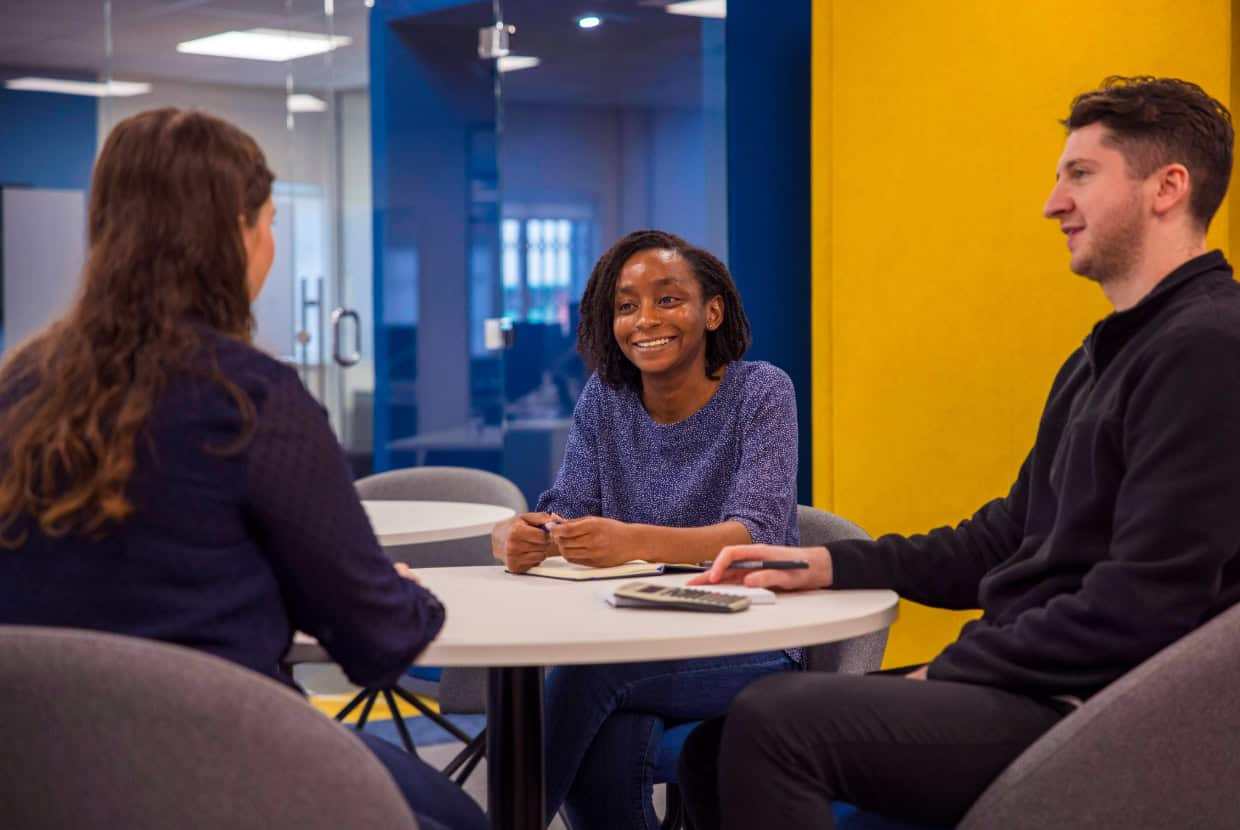 Three people in conversation around a small table in a modern office, with one woman smiling and taking notes