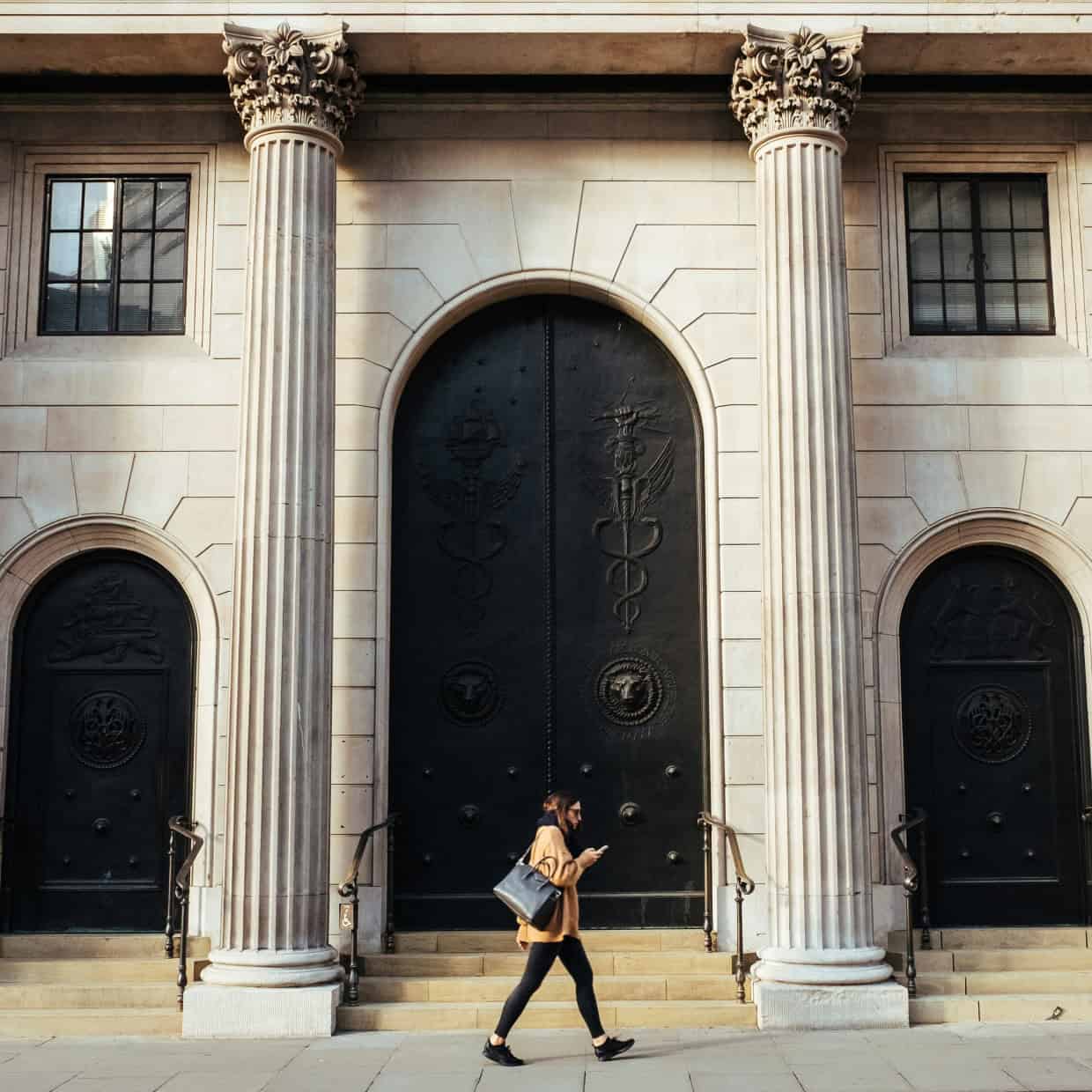 Person walking past a grand neoclassical building with tall columns and large ornate doors, suggesting a historic or financial institution.