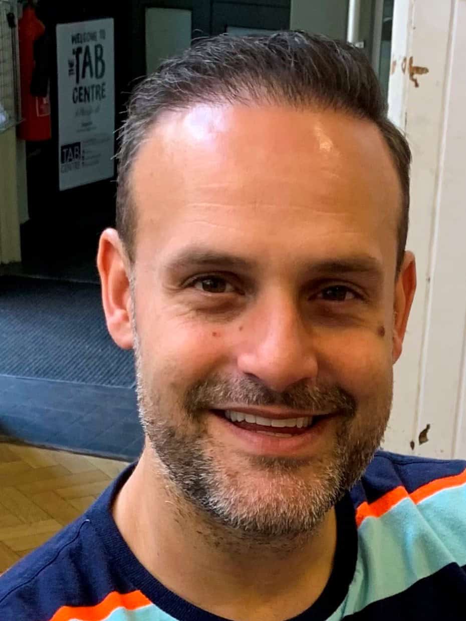 Smiling man with short dark hair and trimmed beard wearing a striped shirt, seated indoors with a sign reading 'The Tab Centre' visible in the background.