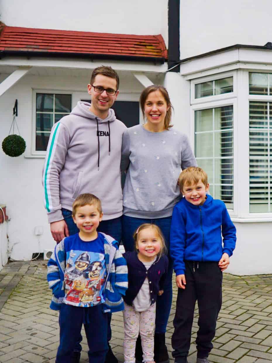 Smiling family of five standing outside their home, with parents behind and three young children in front on a paved driveway.