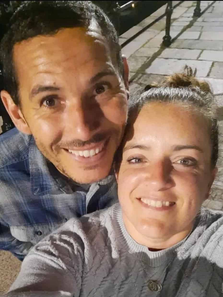 Smiling couple taking a selfie at night on a paved walkway beside water with metal railings in the background.