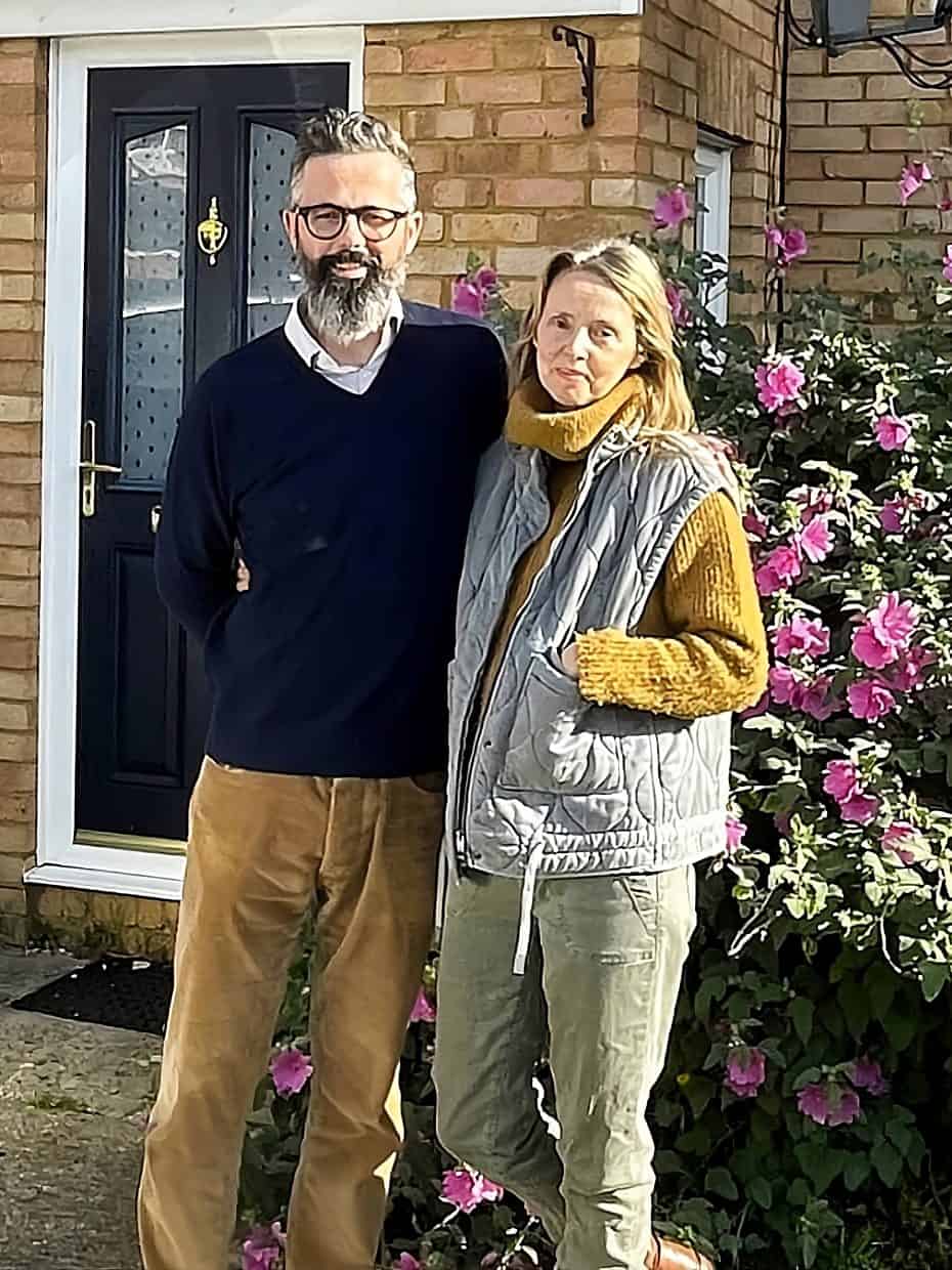 Couple standing together outside a brick house with a black front door, next to a car and a large bush covered in pink flowers